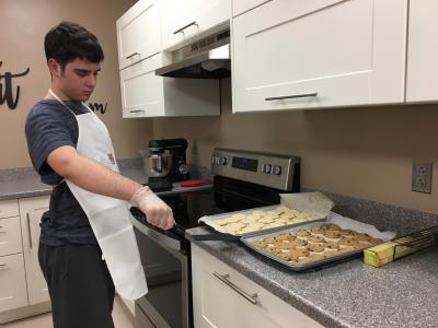 Student standing in kitchen holding a spatula next to 2 trays of baked cookies. 
